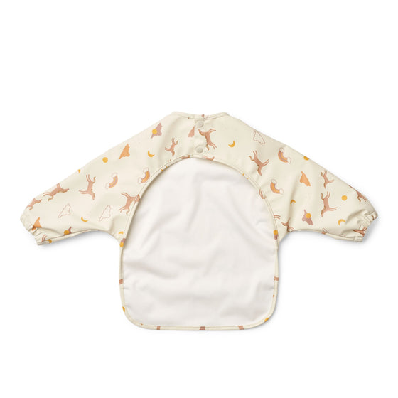 Bavoir à manches longues imperméable pour bébé - Modèle Merle Dream Écru de Liewood, coloris crème avec motifs oniriques : gazelles sautantes, arcs-en-ciel, nuages et soleils. Idéal pour les repas et activités manuelles, facile à nettoyer et doté d’une poche récupératrice.