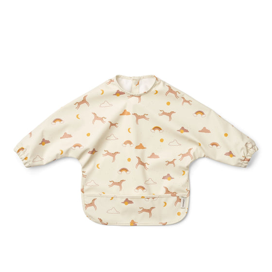 Bavoir à manches longues imperméable pour bébé - Modèle Merle Dream Écru de Liewood, coloris crème avec motifs oniriques : gazelles sautantes, arcs-en-ciel, nuages et soleils. Idéal pour les repas et activités manuelles, facile à nettoyer et doté d’une poche récupératrice.